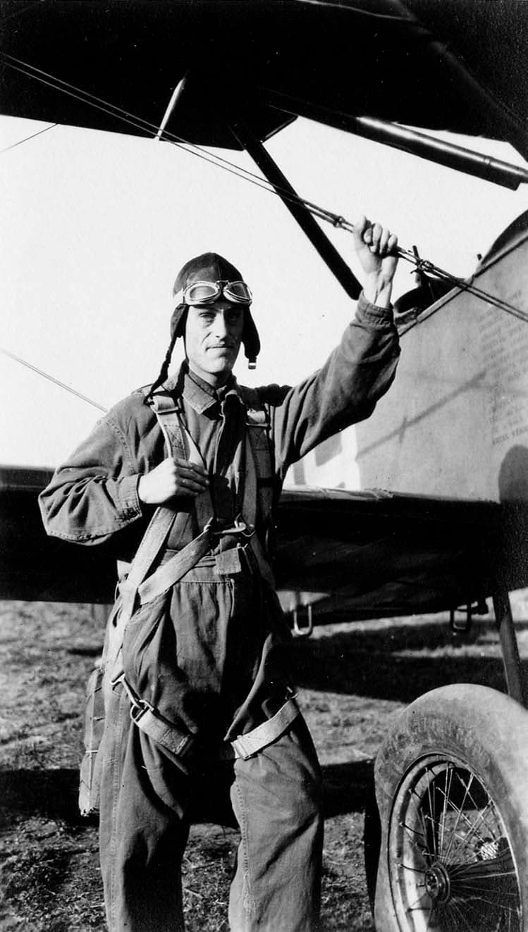 An Army Air Corps flight cadet in a flight suit and leather helmet, holding a bracing wire next to a training biplane.