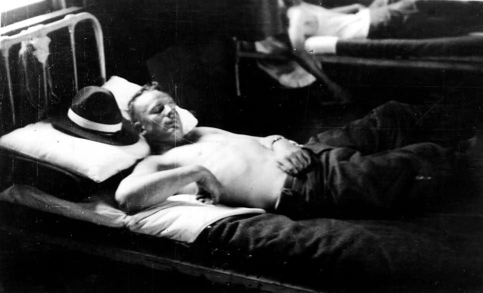 A shirtless flight cadet sleeping deeply on a narrow iron cot in the barracks, with his campaign hat resting on the pillow beside him.