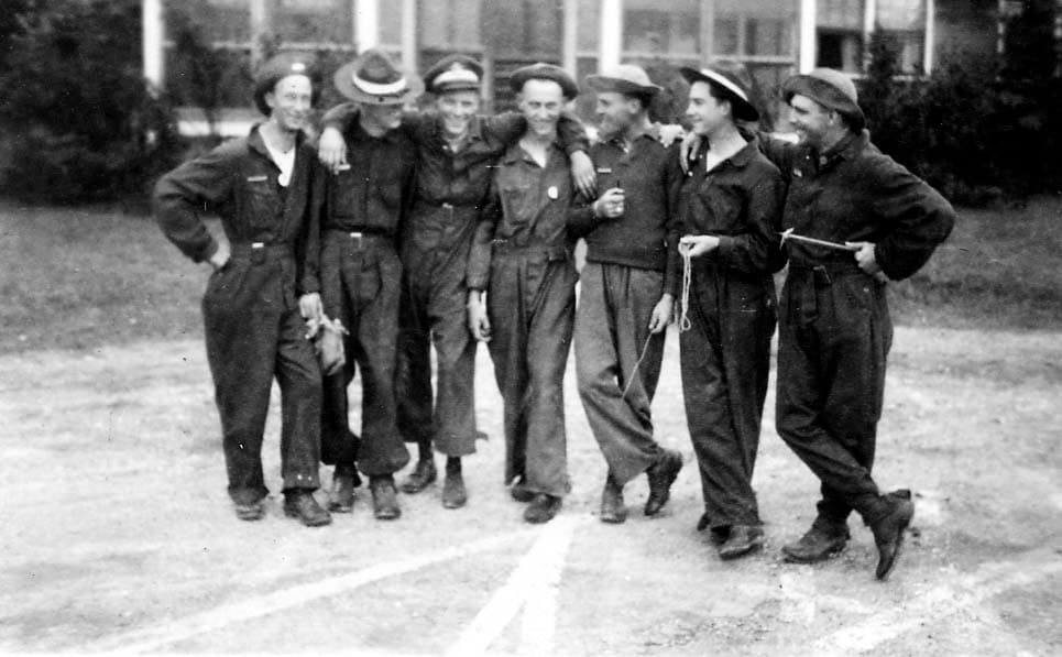 A group of seven smiling flight cadets in dark coveralls and campaign hats standing casually together with their arms around each other's shoulders.