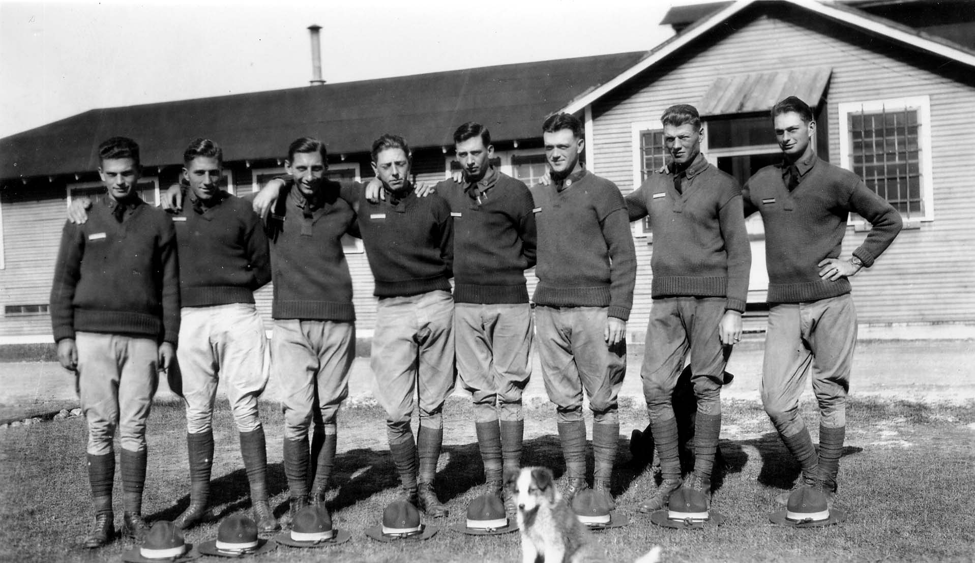 A line of eight smiling flight cadets standing shoulder-to-shoulder in matching sweaters and riding breeches, with their caps and a small dog at their feet.