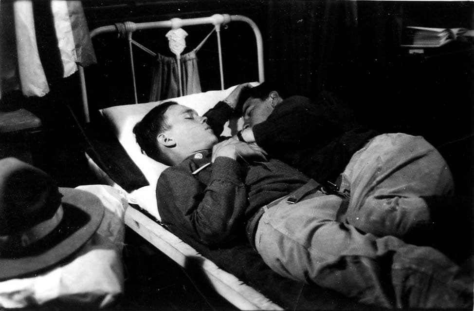 Two fully uniformed flight cadets sleeping closely together on a single narrow cot in the barracks, demonstrating profound exhaustion.