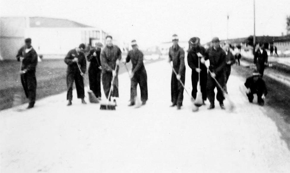 A group of Army Air Corps flight cadets in uniform sweeping a wide dirt or gravel road with push brooms.