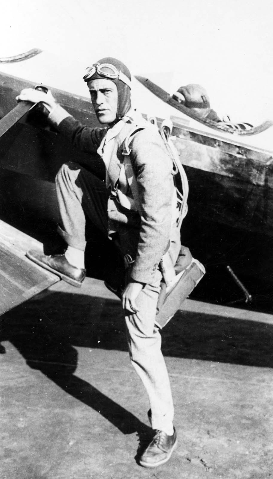 Donald A. Hall in leather aviator cap, goggles, and parachute harness climbing into an airplane cockpit.