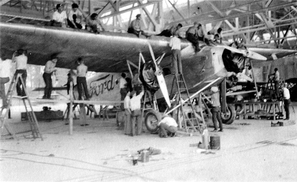 A large crew of men working on the massive, corrugated metal wing and fuselage of a Ford Tri-Motor inside a brightly lit hangar.