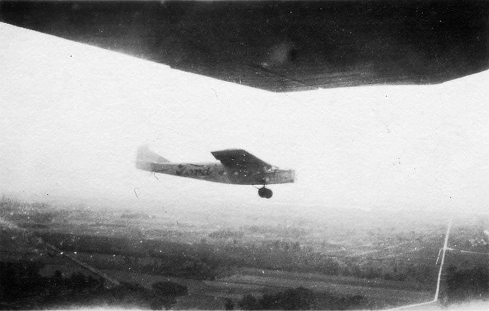 An air-to-air photograph taken from beneath the wing of another aircraft, showing a Ford Tri-Motor passenger plane flying over a rural landscape.