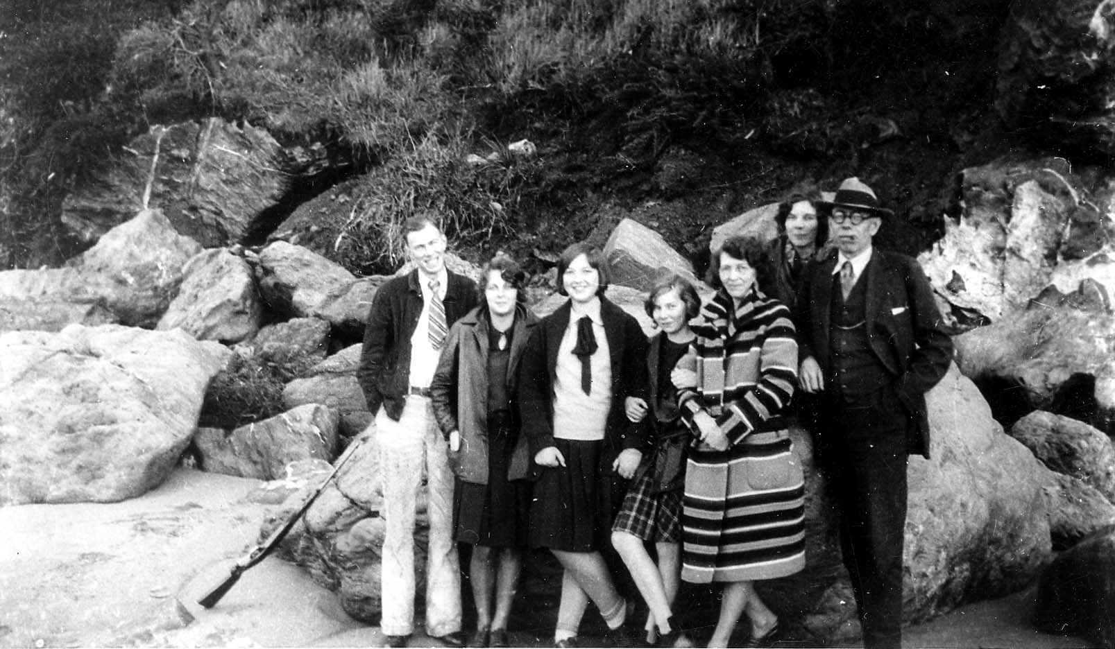 A group of six friends, bundled in coats and jackets, standing together on a sandy beach at the base of a rocky, cliffside bluff.