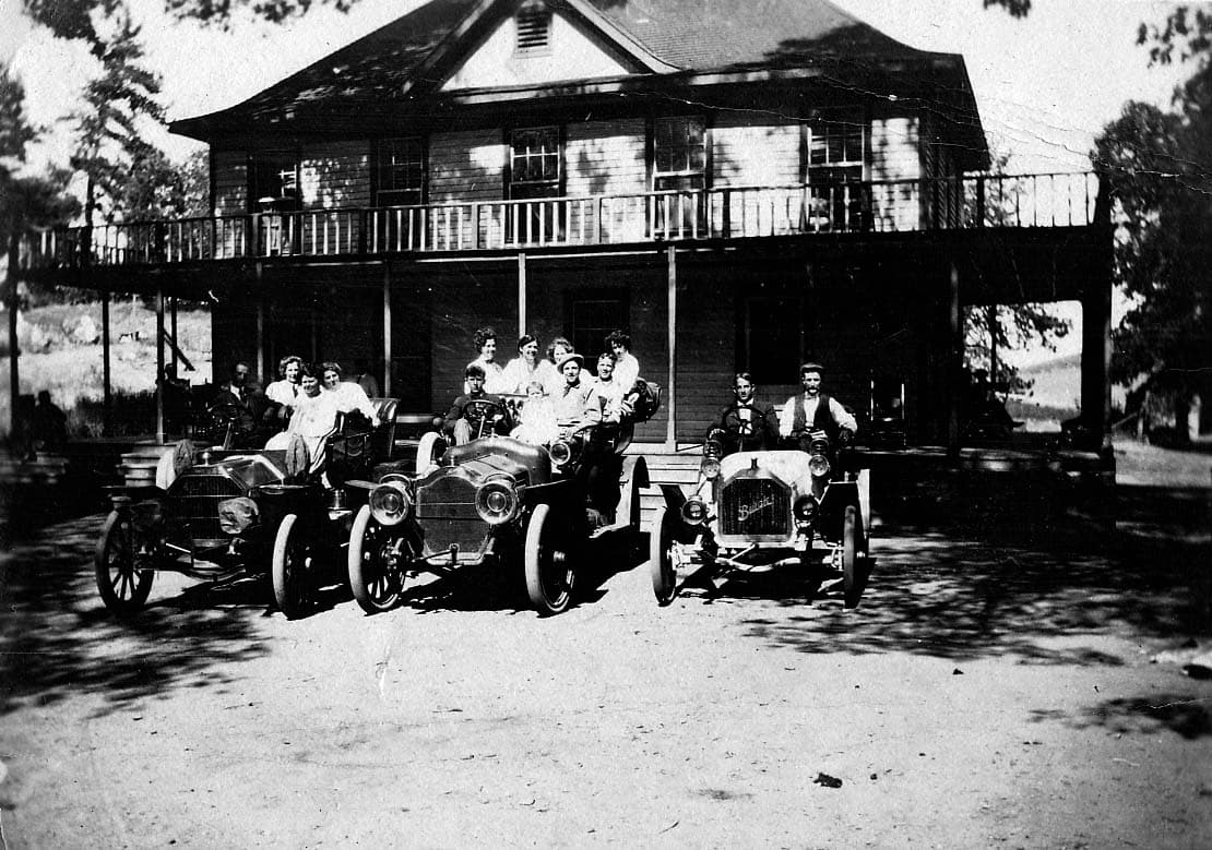 A large group of friends posing in and around three vintage open-cab automobiles parked in front of a two-story wooden lodge.
