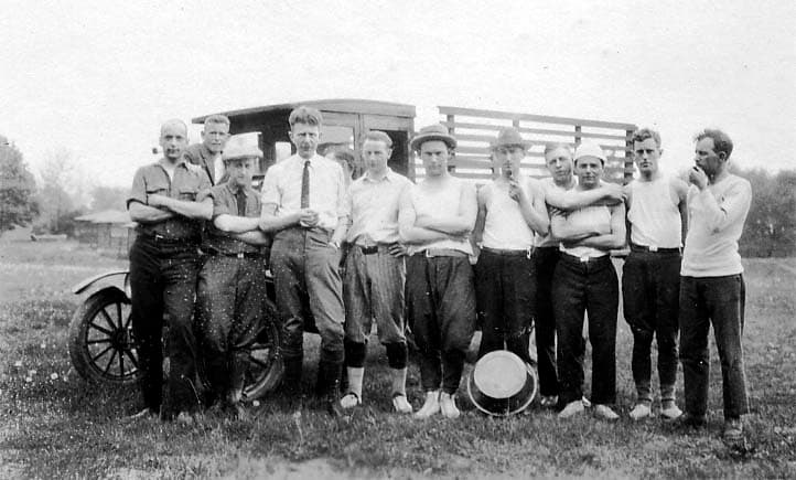 A large group of men, many in undershirts or work clothes, posing proudly in a grassy field in front of an early flatbed truck.