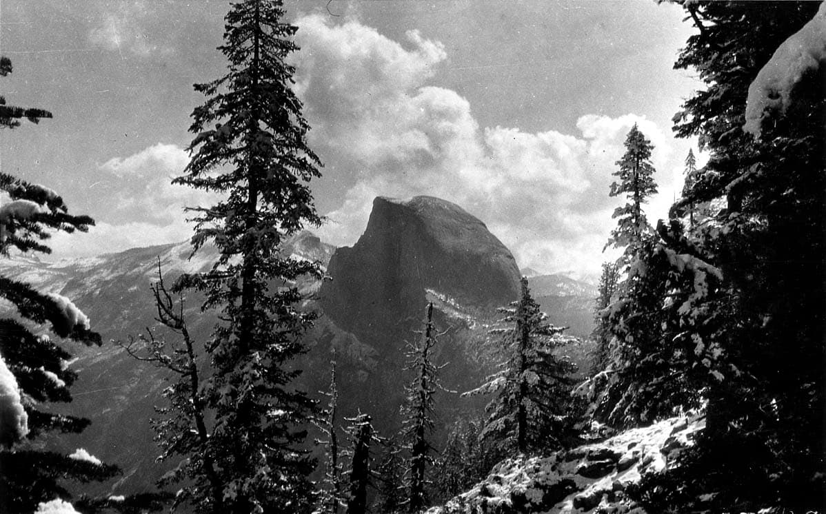 The sheer granite monolith of Half Dome rising in the distance, framed by snow-covered conifers under billowing cumulus clouds.