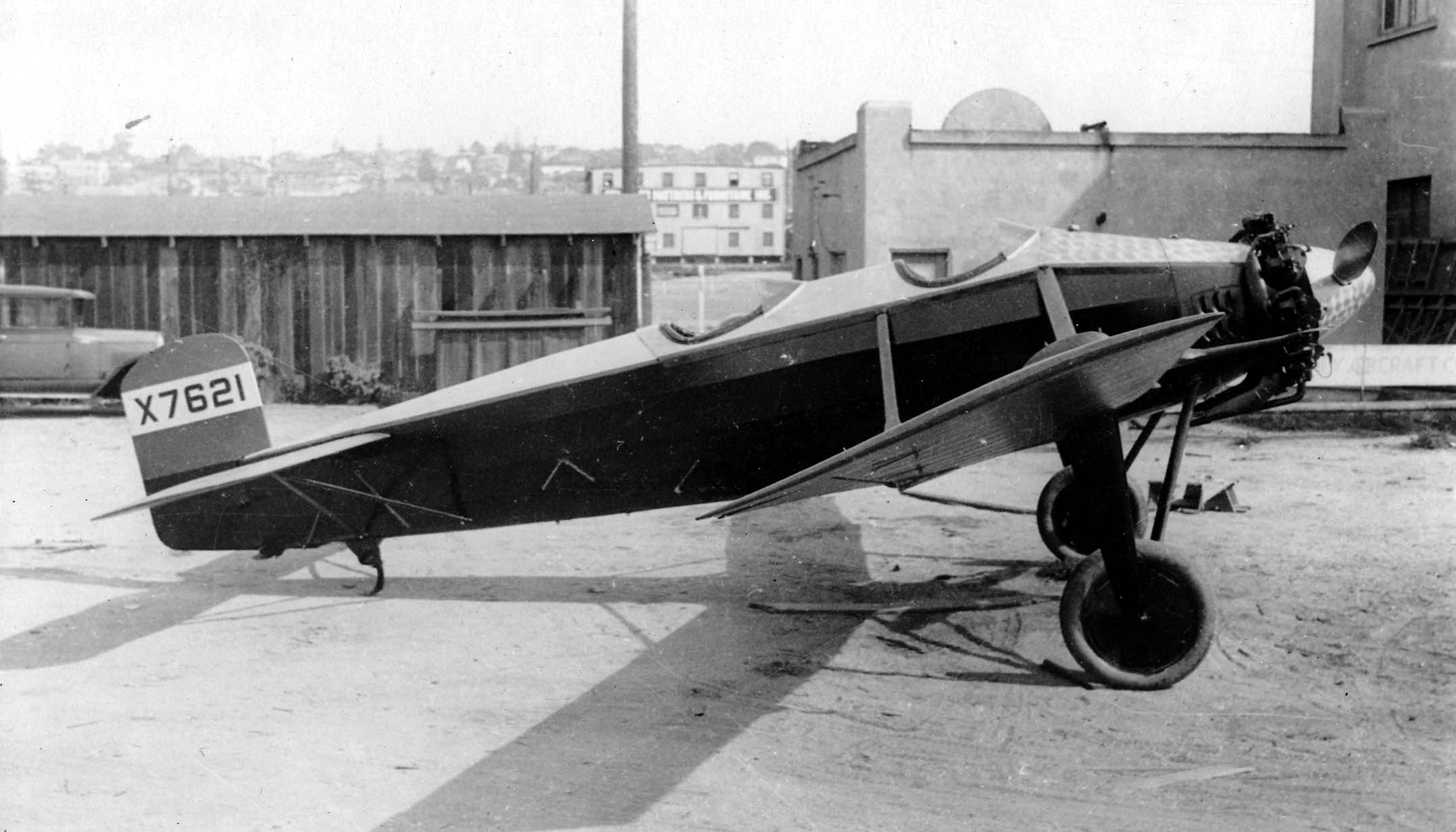 A crisp side profile of the experimental Hall X-1 tandem-wing monoplane X7621 with an engine-turned nose cowl parked outside a San Diego factory building.