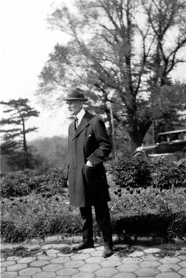 A full-length portrait of Harland Hall in an overcoat and fedora standing on a hexagonal stone pathway in a formal park garden with a vintage car in the background.