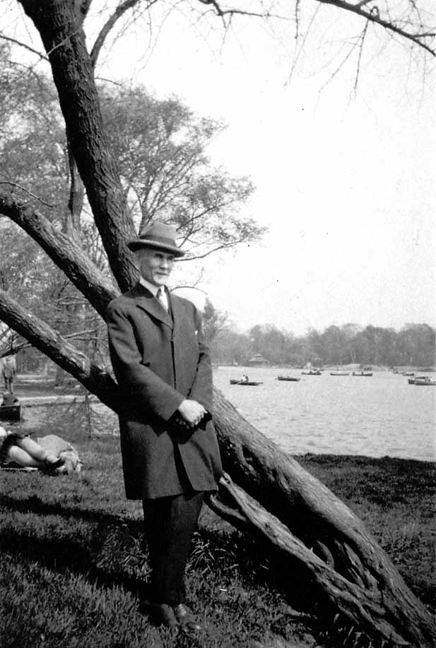 A full-length portrait of Harland Hall in a dark overcoat and fedora standing beside a leaning tree near a public park lake with rowboats.
