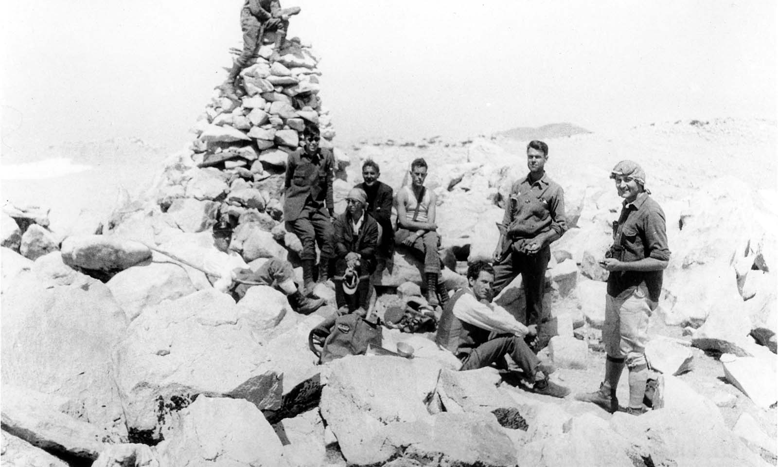 A group of hikers resting among large rocks next to a tall stone cairn at a mountain summit.