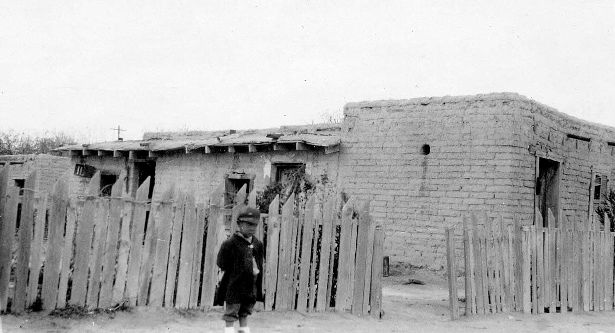 A weathered, flat-roofed adobe house with a crooked wooden fence and a child standing in the foreground.