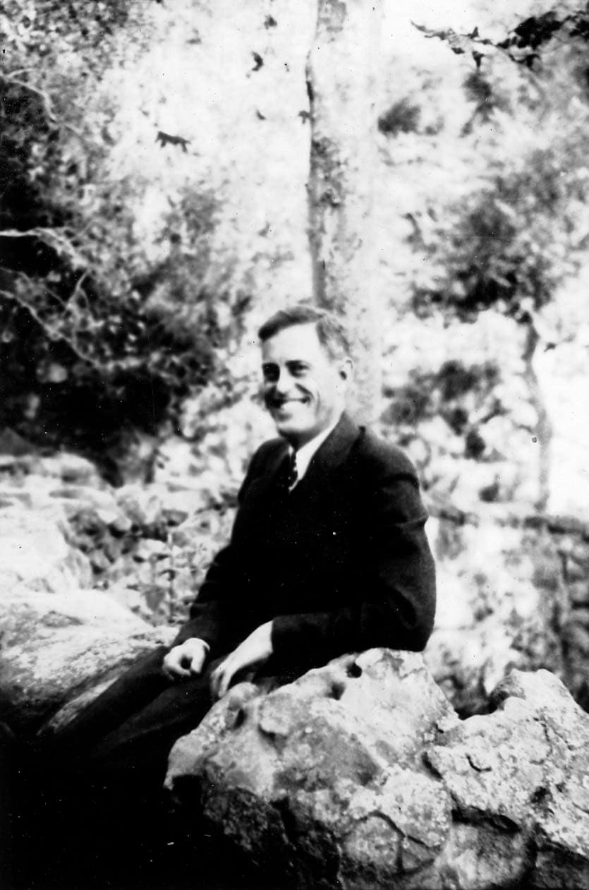 A portrait of Donald A. Hall Sr. smiling broadly, wearing a dark suit, sitting on the same rustic stone wall in a wooded area.