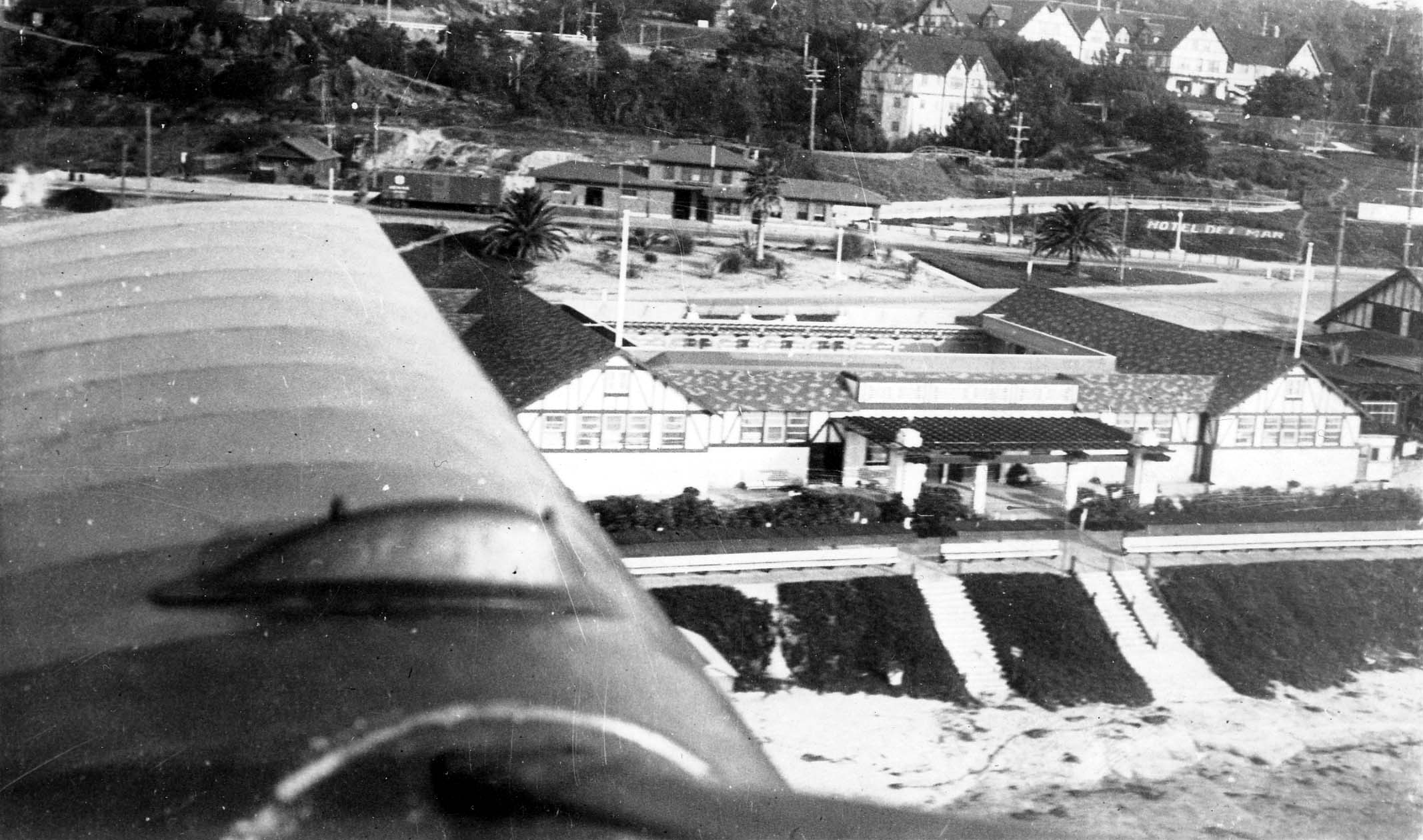 An aerial view looking over an aircraft wing down at the historic Tudor-style Hotel Del Mar, coastal railroad tracks, and ocean bluffs.