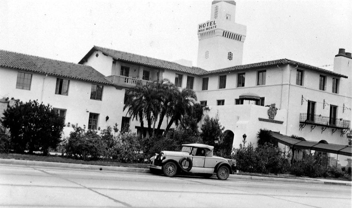 An exterior view of the Hotel Mar Monte with a vintage, two-door convertible automobile parked on the street in front of the Spanish-style arches.