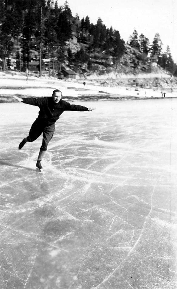 An action photograph of a man ice skating on a frozen lake, balancing on one foot with his arms outstretched, with a snowy, wooded hill in the background.