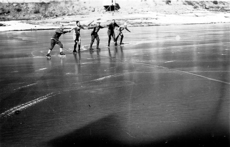 A group of four men ice skating together in a line, holding hands as they glide across the frozen, expansive surface of a lake.