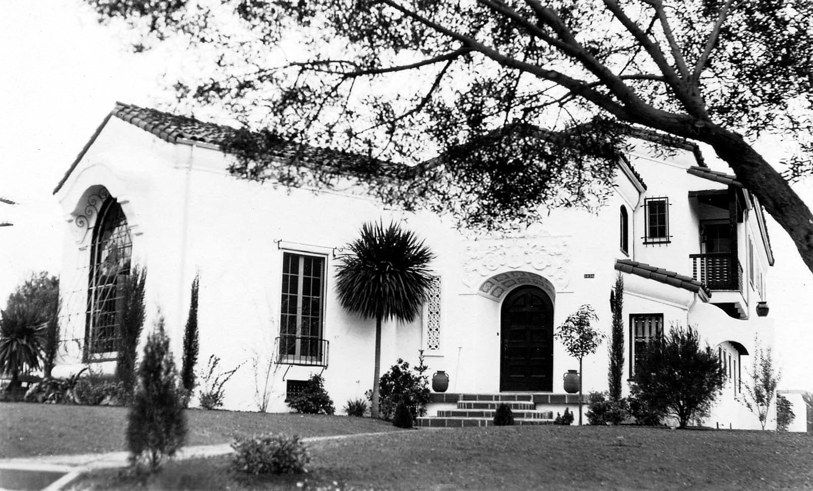 A wide exterior shot of a large, wealthy estate featuring Spanish Colonial Revival architecture, a manicured lawn, and a prominent arched entryway.