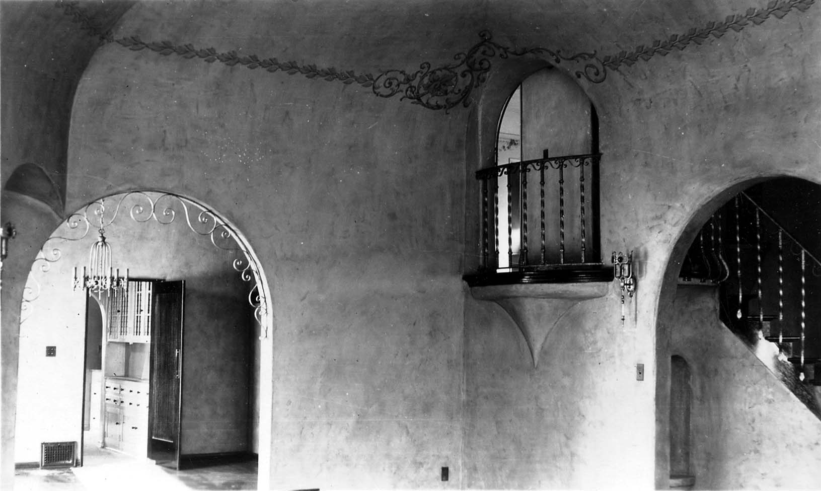A highly detailed interior shot showing an arched doorway, an ornate wrought-iron balcony, and a sweeping staircase inside the Spanish-style estate.