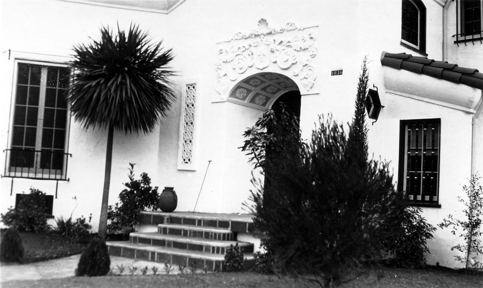 A close-up architectural view of the ornate, carved entryway and tiled steps of a wealthy Spanish Colonial Revival home, flanked by a large palm tree.