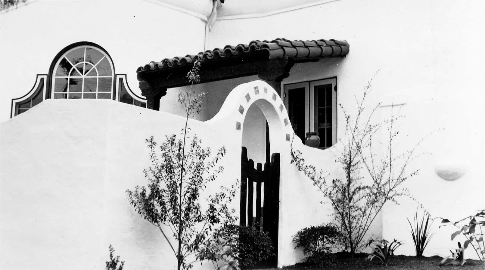 An exterior architectural view looking through a white stucco archway toward a dark wooden door and a tiled patio with potted plants.