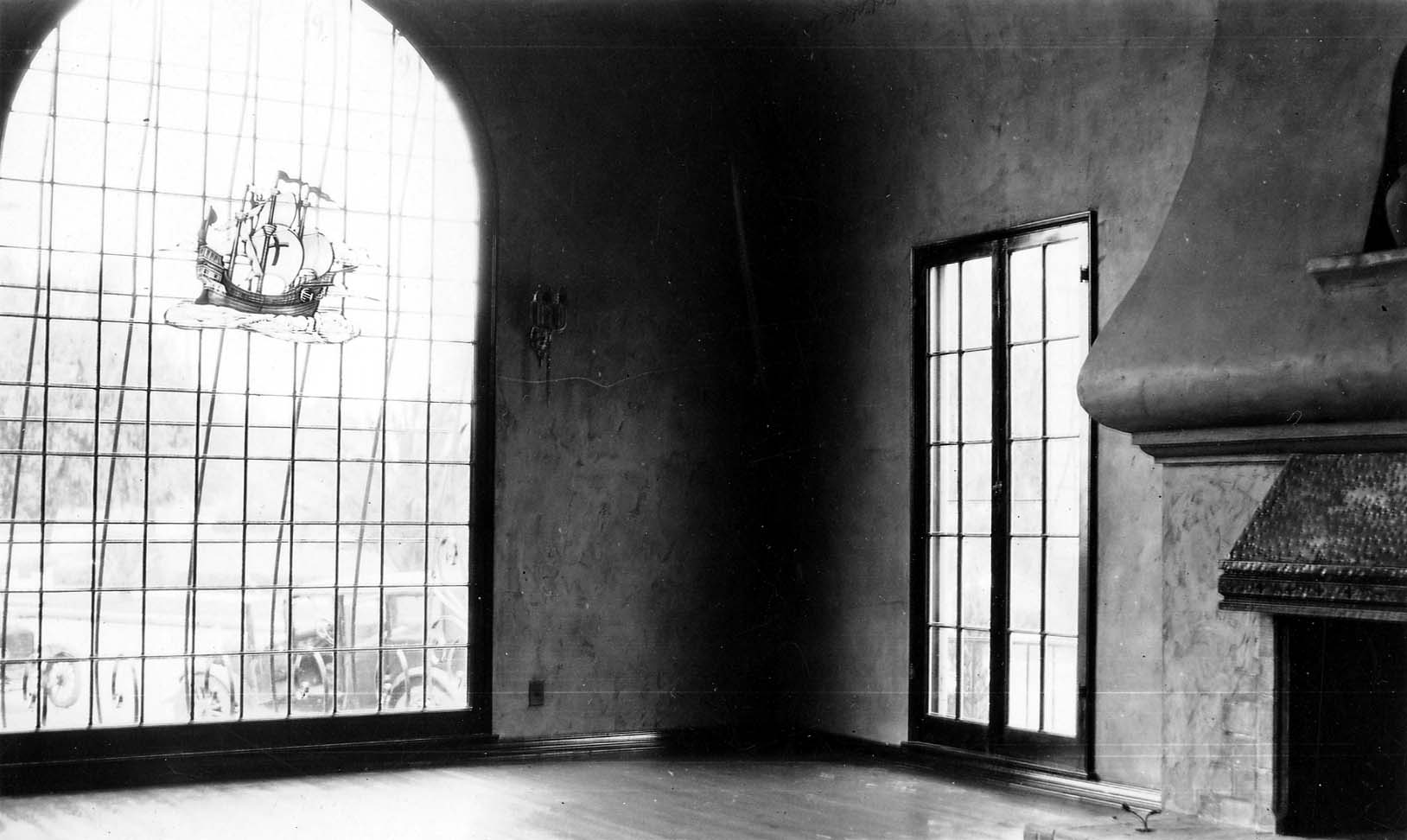 An interior view of a large, dark room featuring a massive stained-glass window with a ship motif next to a large stucco fireplace.