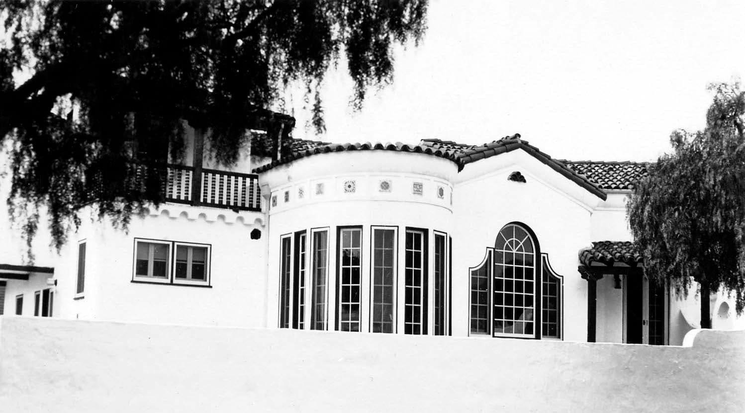 An exterior architectural shot highlighting the rounded turret, multi-paned windows, and a dark wooden balcony of a large Spanish-style estate.