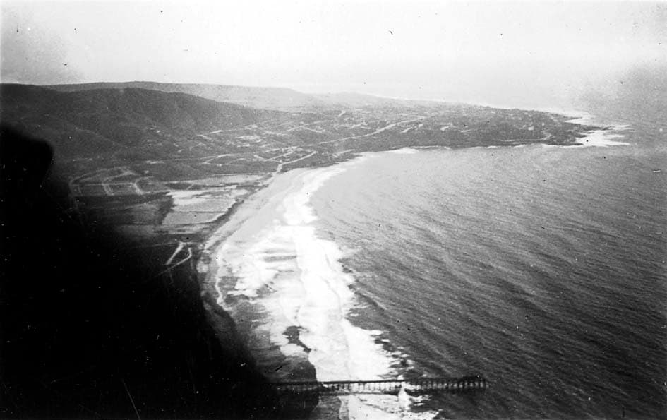 An aerial perspective looking south along the San Diego coastline, showing the Scripps Institution of Oceanography Pier, the crescent of La Jolla Shores, and La Jolla Cove.