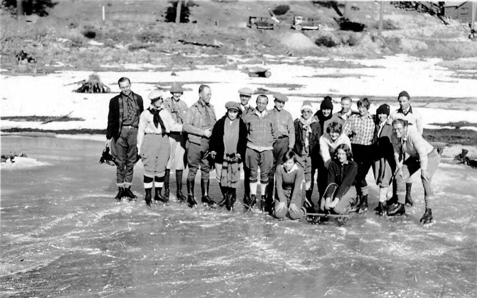 A large, mixed group of men and women wearing winter gear and ice skates, posing together for a group portrait on a frozen lake.