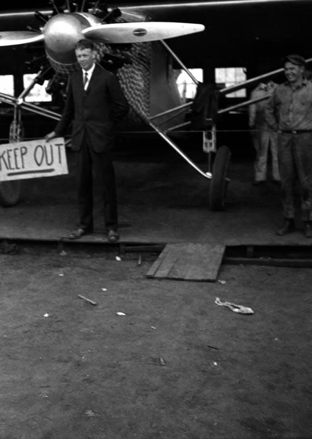 Charles Lindbergh standing near the wing of the Spirit of St. Louis, holding a hand-painted "KEEP OUT" sign.