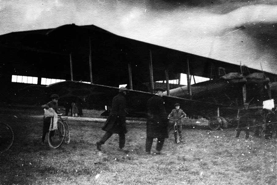The giant LWF Model H Owl three-engine biplane bomber parked outside a hangar with onlookers and boys on bicycles in the foreground.