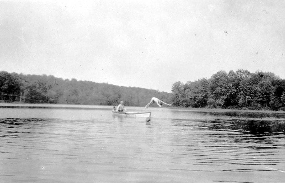 A man diving headfirst into a lake from the edge of a small wooden rowboat, while another man sits in the boat watching.