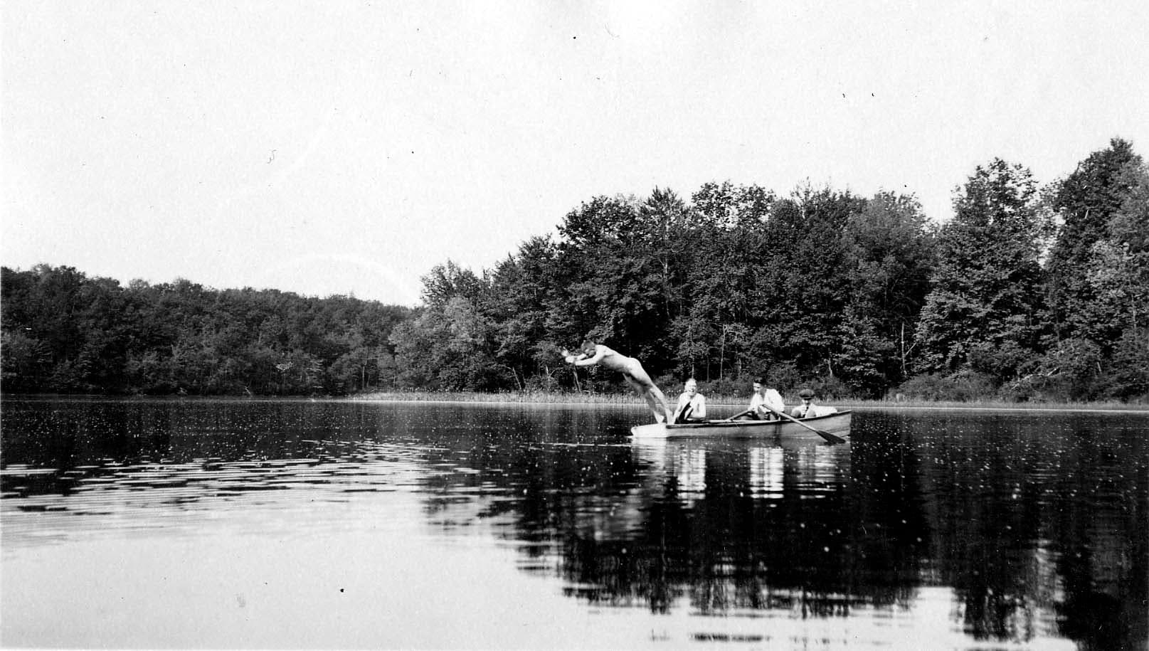 A man diving headfirst from a wooden rowboat into a calm, tree-lined lake, while two other men sit in the boat holding the oars.