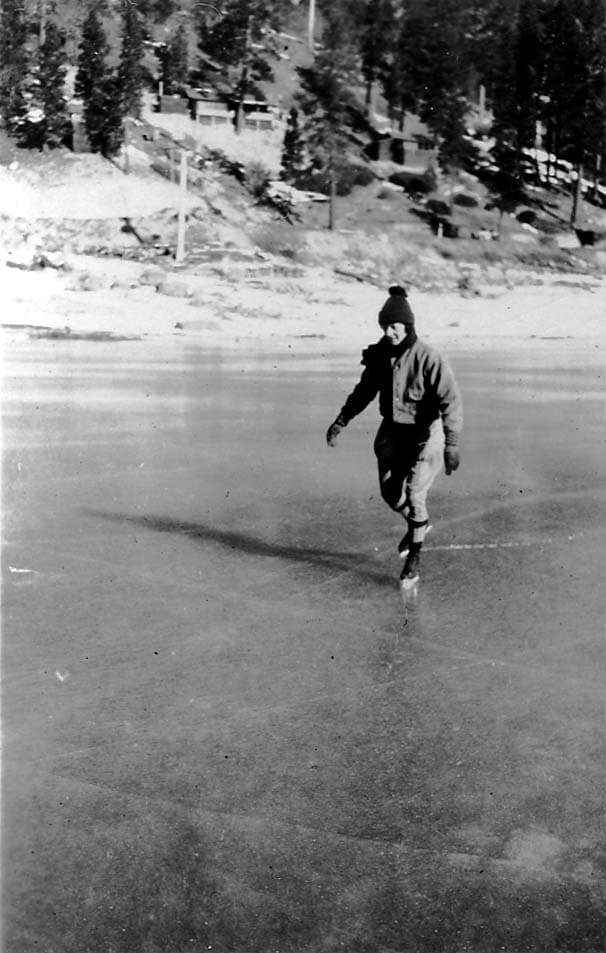 A man wearing a knit cap and a heavy jacket ice skating gracefully across a frozen lake, with a snowy, wooded hillside in the background.