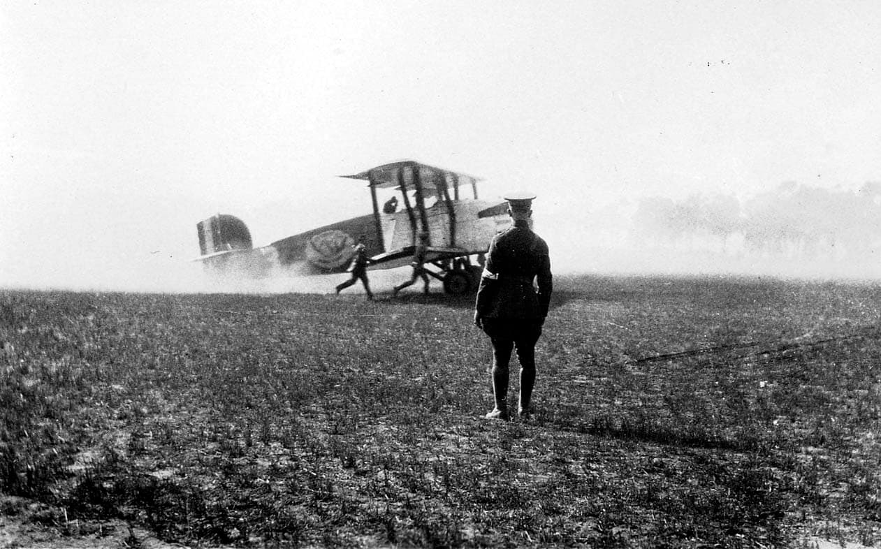 A military biplane kicking up dust on a tarmac as crew members rush toward it, watched by a military officer in uniform.