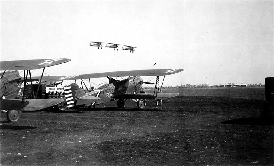 A low-angle view of three military biplanes flying in formation over several parked aircraft on a dirt airfield.