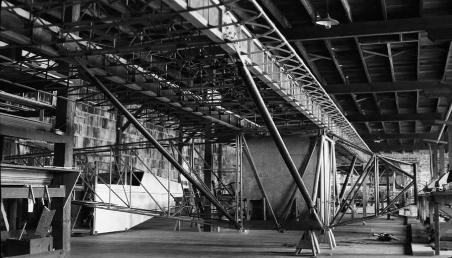 The raw, uncovered tubular steel framework of an aircraft fuselage sitting inside a wooden hangar.