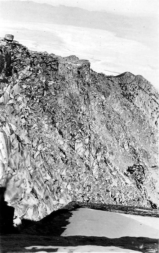 A high-altitude view looking down a steep, rocky mountain cliff face toward a distant valley and dry lakebed in Southern California.