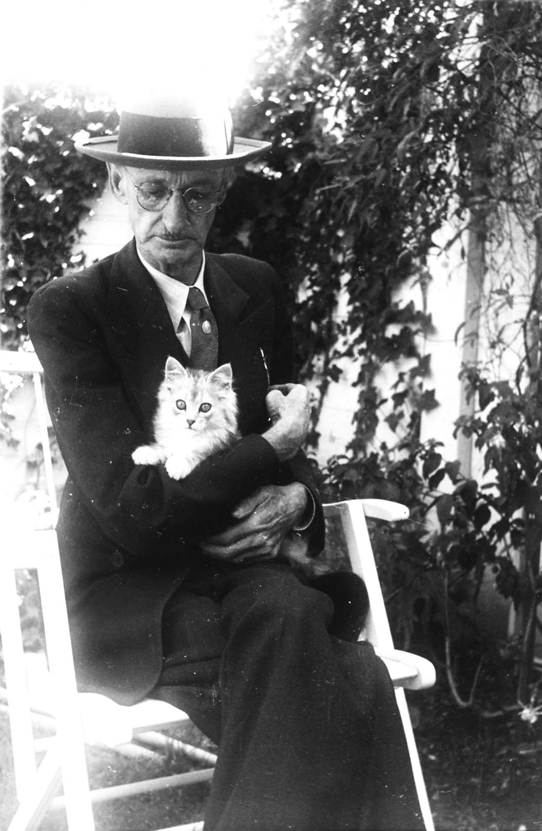 A portrait of an Hall Family member of older gentleman wearing a dark suit, tie, and fedora, sitting in a white chair and holding a fluffy white kitten.