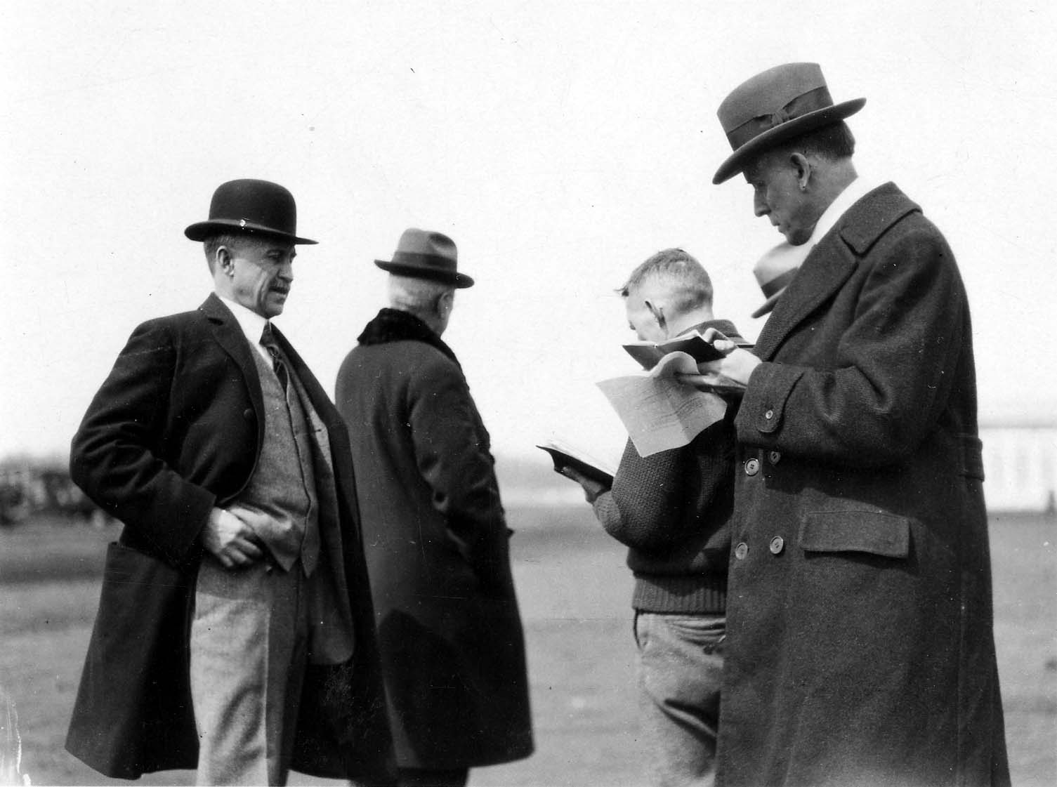 A historic photograph of Orville Wright in a bowler hat and overcoat conversing with aviation officials and journalists recording notes on an airfield.