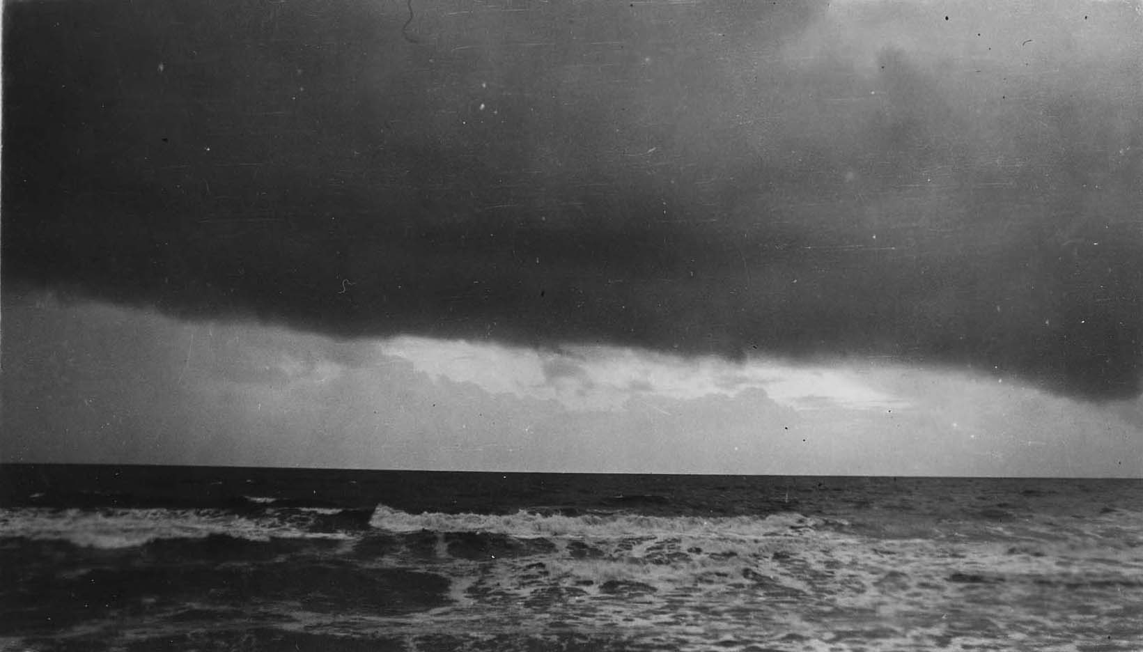 A heavy, dark squall cloud hanging low over a flat ocean horizon with foaming white surf breaking along the shoreline.