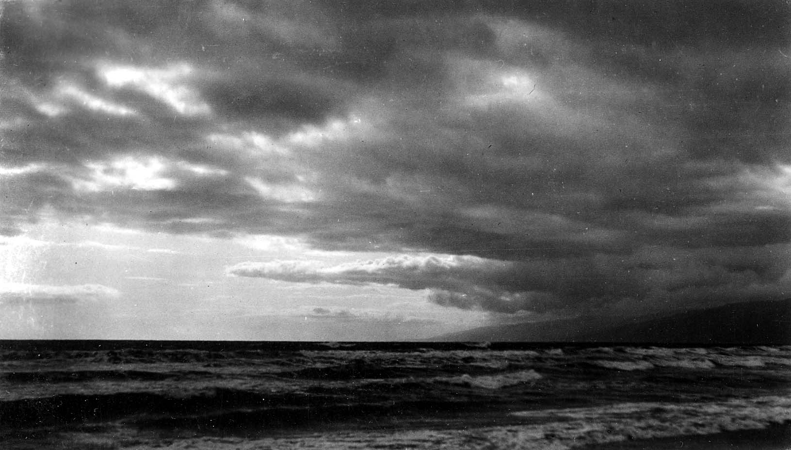 Choppy ocean waves and whitecaps rolling toward a sandy shore beneath turbulent, wind-swept clouds and a distant coastal headland.