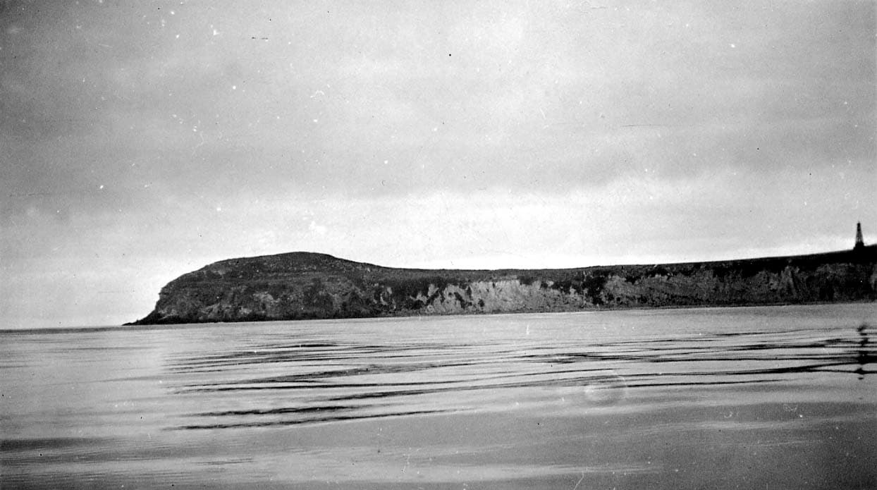 A low-angle maritime perspective of a flat-topped coastal promontory and sheer cliffs meeting calm ocean waters under an overcast sky.