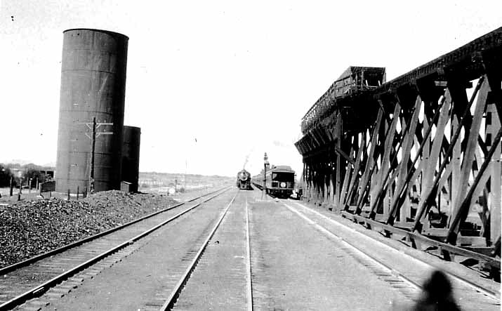 A steam train in the distance passing between large cylindrical water towers and a massive wooden industrial loading trestle.