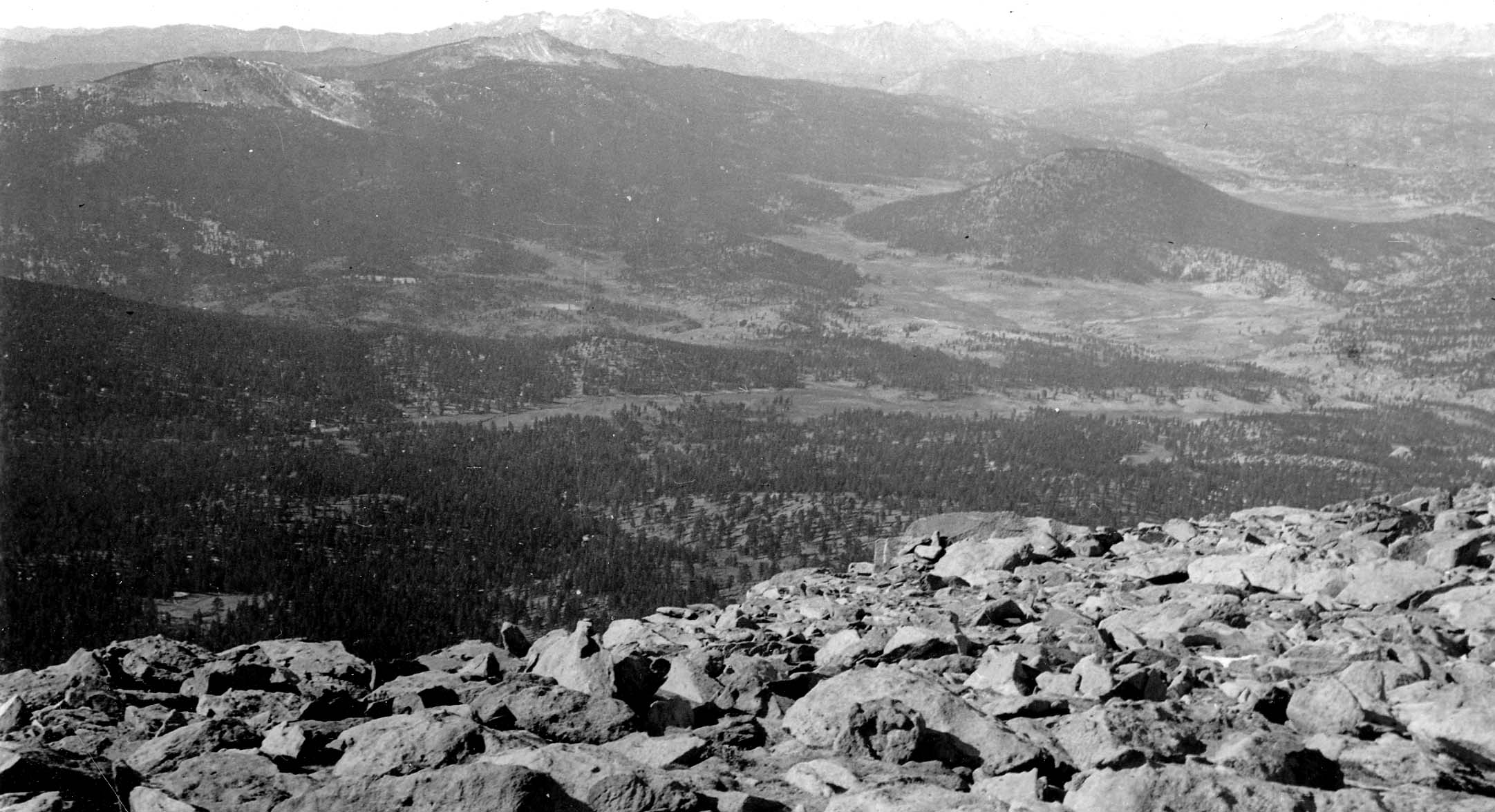 A sweeping high-altitude view from a boulder-strewn summit looking out over a wide forested valley with a distinct volcanic cinder cone in the midground.