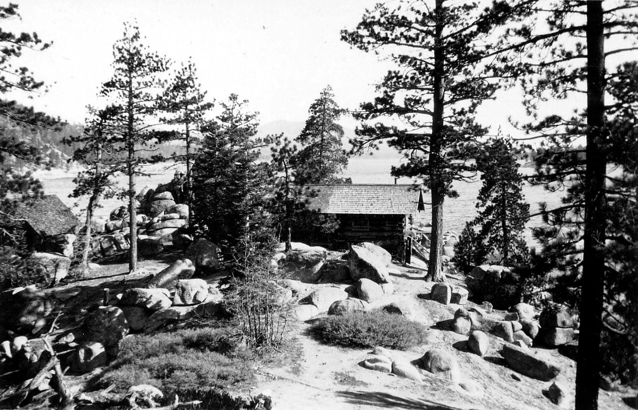 A rustic log cabin nestled among massive granitic boulders and tall pine trees on the rugged shore of a mountain lake.