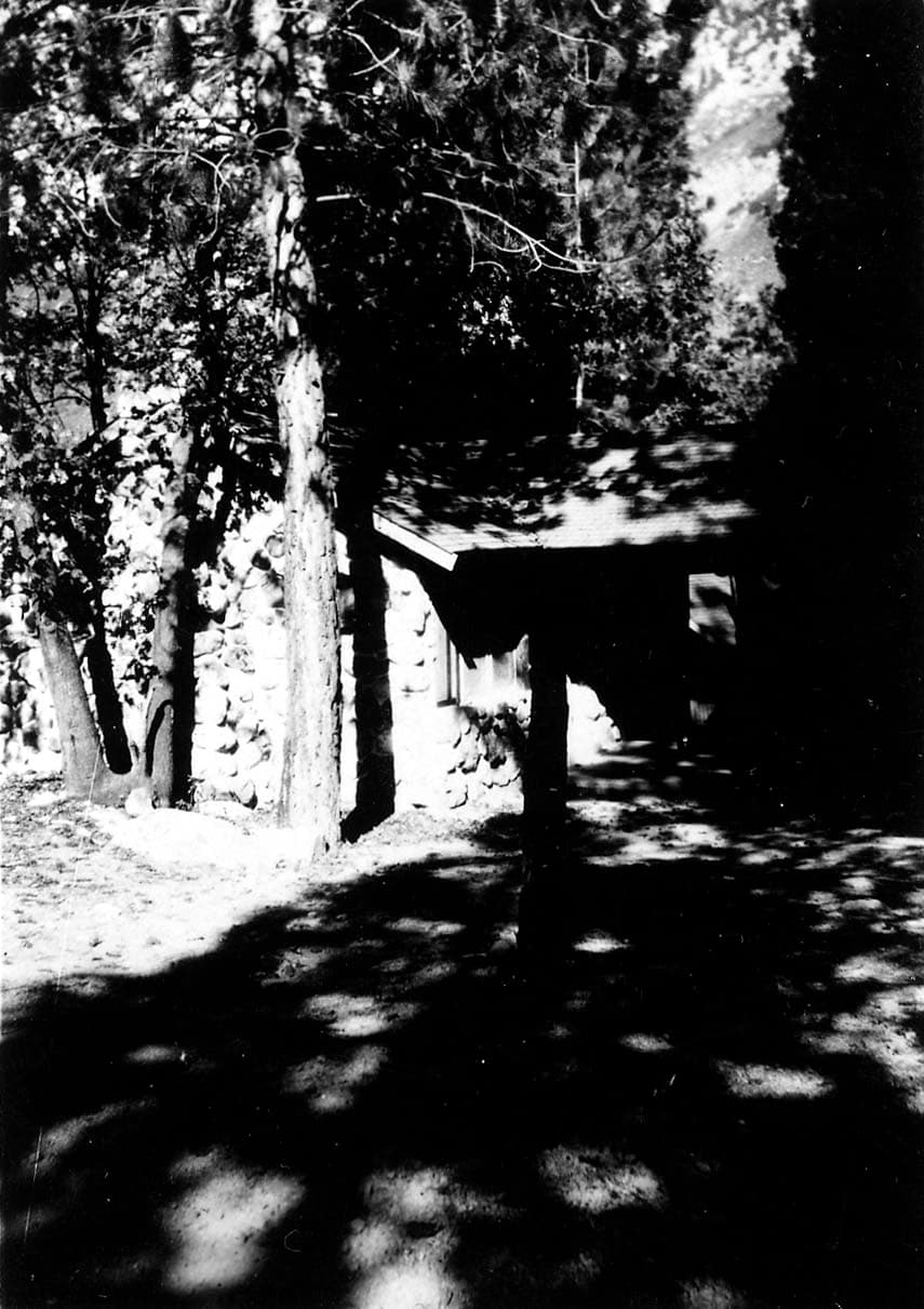 A rustic stone and timber mountain cabin nestled in dappled shade among tall pine trees in the San Bernardino Mountains.