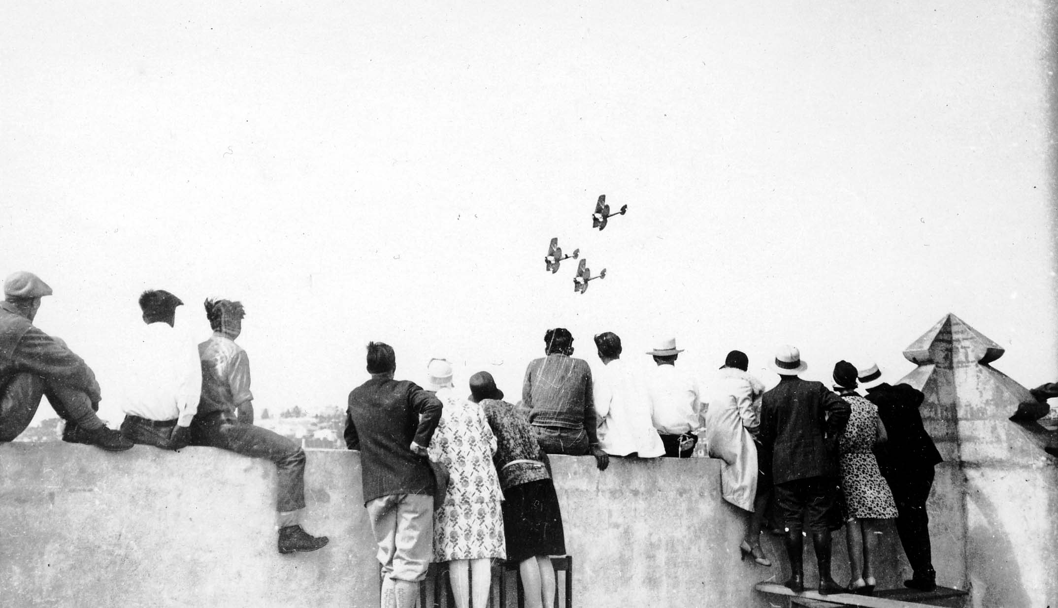 A row of men and women, likely Ryan Airlines employees, sitting and standing on the edge of a factory roof, watching three biplanes dive in the distance.